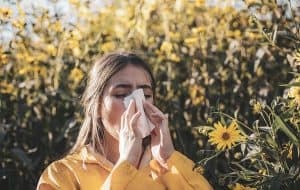 Young woman blowing her nose and experiencing seasonal allergies while outdoors in a field of yellow flowers