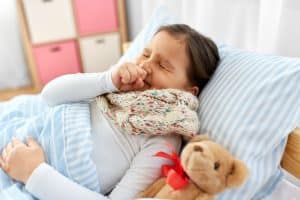 Picture of a young girl with RSV lying in bed next to a teddy bear and coughing.