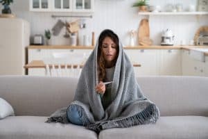 Picture of a woman with an upper respiratory infection sitting on a couch, under a blanket, and checking a thermometer.