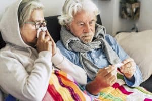 Picture of a sick older couple sitting on a couch. The woman is blowing her nose into a tissue, and the man is holding a thermometer.