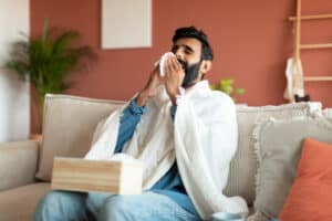 Picture of a man sneezing into a tissue as he sits on a couch with a blanket wrapped around his shoulders.