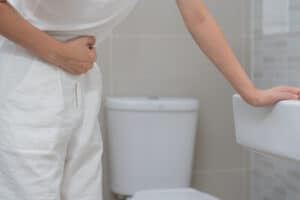Picture of a man with a stomach ache holding his abdomen in pain while standing next to a bathroom sink with a toilet in the background.