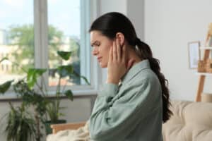 Picture of a woman with an ear infection sitting on a couch and touching her left ear in pain.